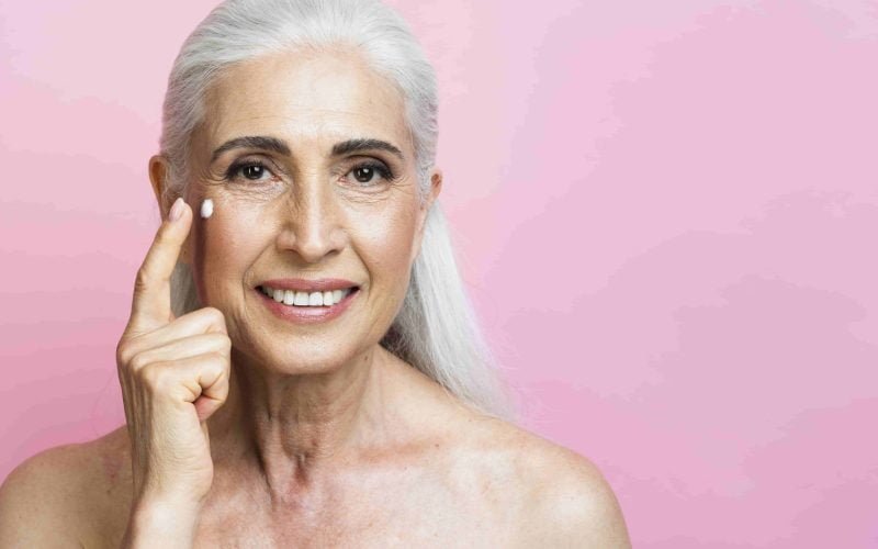 Smiling mature woman applying anti-aging moisturizer to highlight face care essentials.