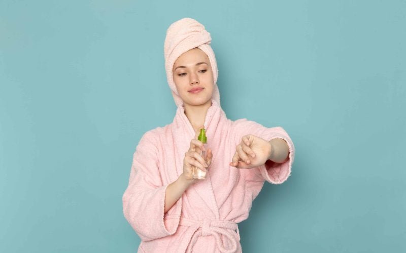 Woman in a pink bathrobe using a body spray, evaluating the safety of your daily products.