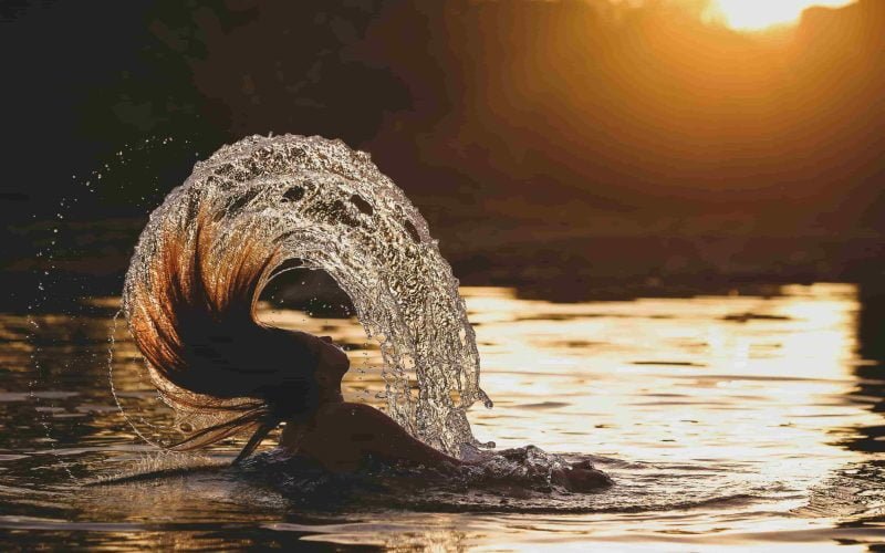 Hair care during different seasons with a woman flipping her wet hair in the water at sunset